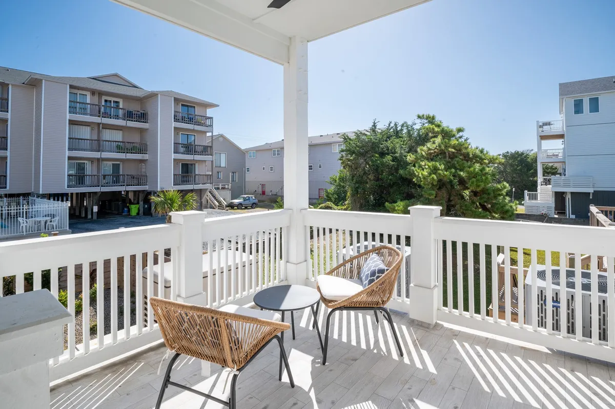 A cozy balcony furnished with wicker chairs and a small round table is surrounded by lush green foliage and nearby coastal buildings. The setting is casual and private, ideal for enjoying a quiet morning or winding down after a day at the beach with a pet nearby.