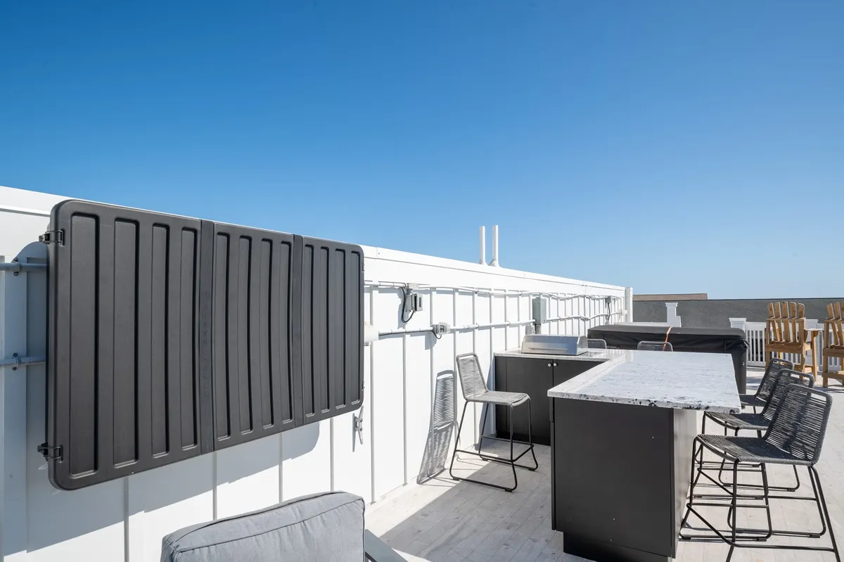 An expansive rooftop deck with a modern bar, outdoor seating, and a clear blue sky overhead. The arrangement is perfect for al fresco dining, sunbathing, and group celebrations, with ample space for everyone.
