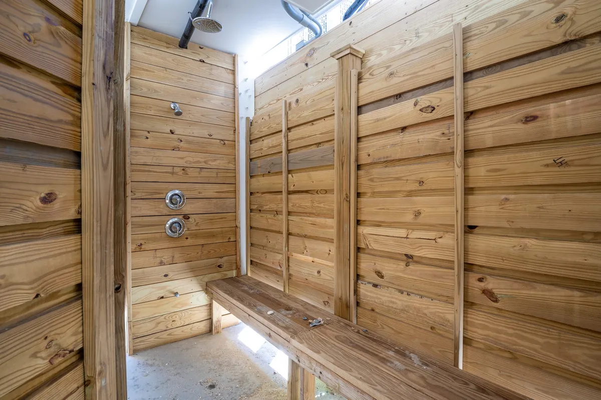 A welcoming outdoor shower area constructed with wooden walls and shower fixtures, featuring a wooden bench for convenience. The design merges rustic charm with practicality, set in a natural and airy space perfect for rinsing off after a day at the beach.