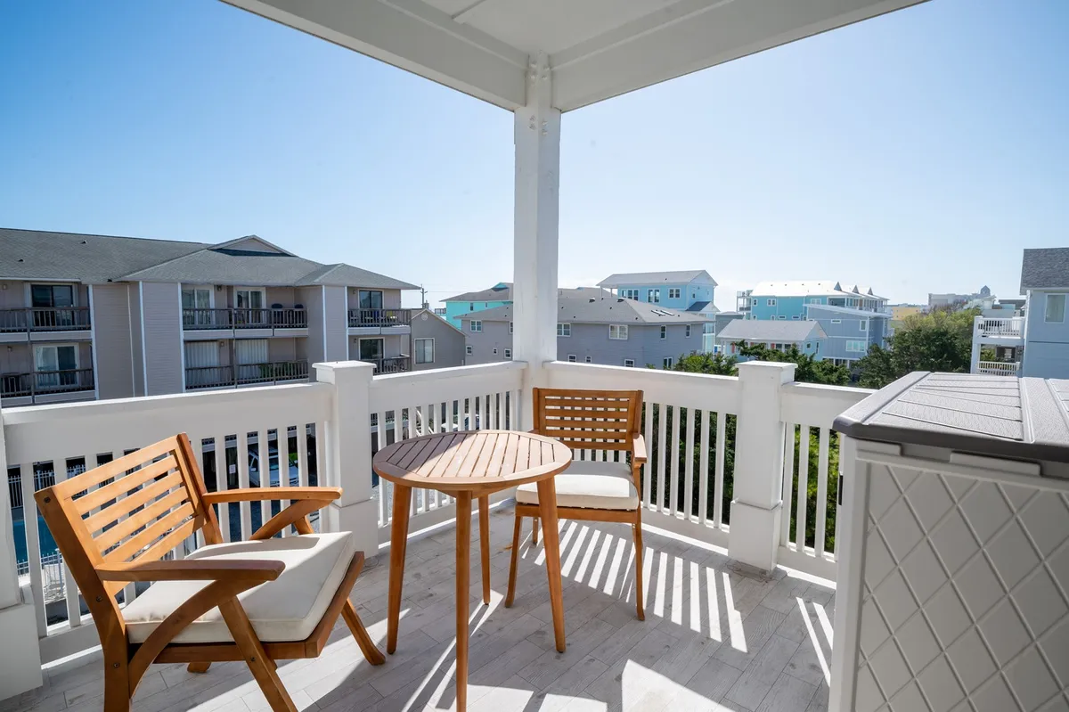 An inviting outdoor porch setup with wooden chairs and a round table overlooks the surrounding coastal community. The space is open and airy, offering a relaxing spot with a view of adjacent buildings, perfect for enjoying a morning coffee or spending time outside with pets.