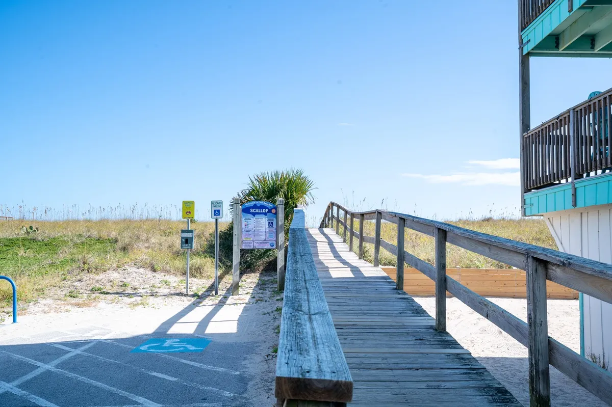 A casual, sunlit wooden walkway leads through palm trees and grass, ending at a welcoming beach access sign. The scene feels relaxed and inviting, inviting visitors to walk or bike straight onto the sandy shore just steps away.
