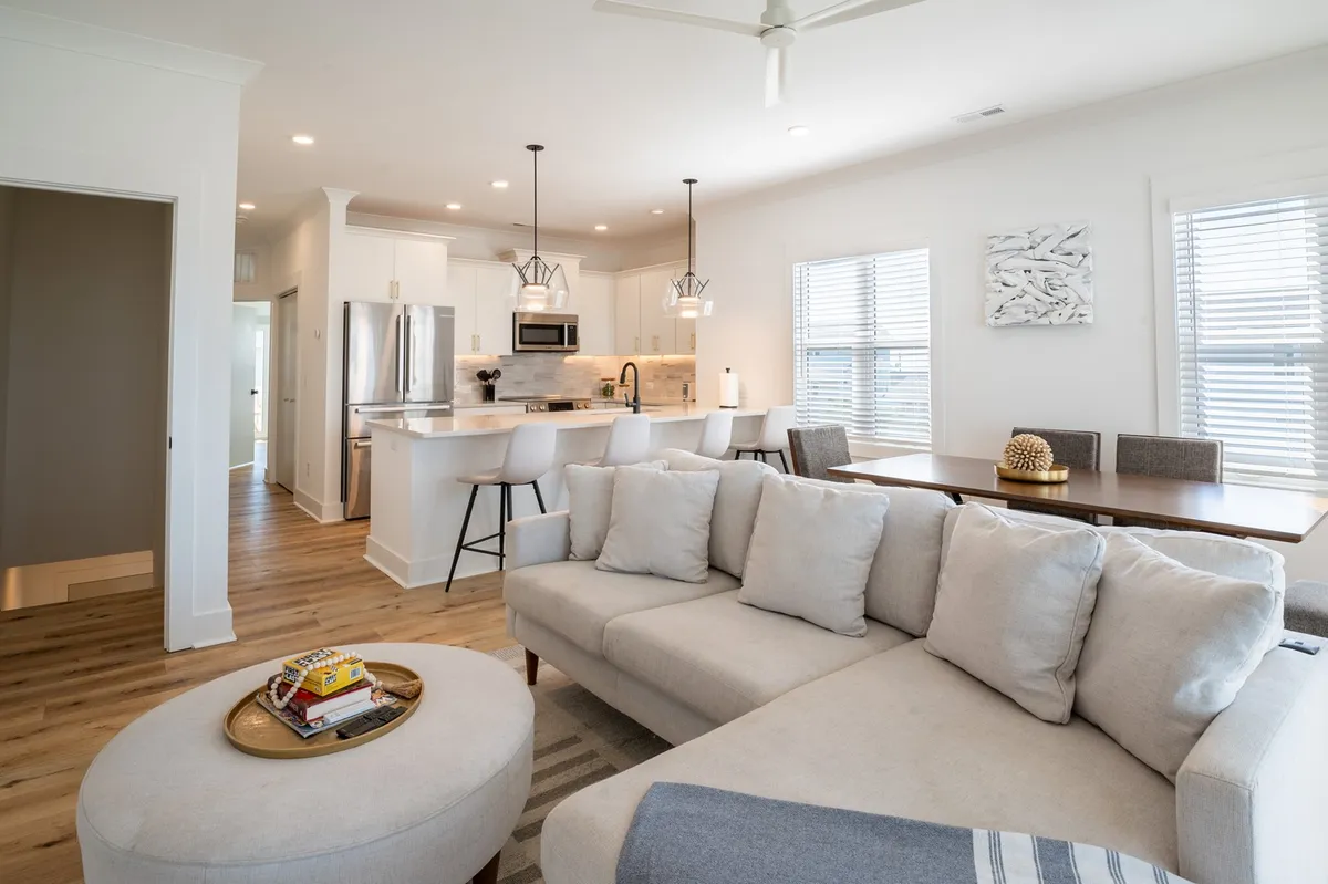 A spacious, open-plan interior featuring a comfortable sofa, a dining table set for gatherings, a large kitchen island, and warm lighting fixtures. The area is bathed in natural light, inviting guests to socialize, share meals, or relax together.