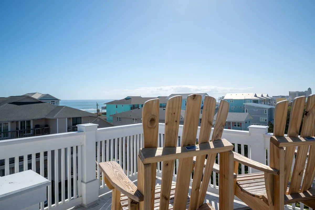 Two wooden Adirondack chairs are positioned on a raised deck, facing a sweeping ocean view beneath a bright blue sky. This tranquil scene invites moments of relaxation, socialization, and sunset-watching—perfect for guests and their pets to enjoy the seaside atmosphere.